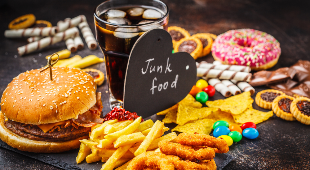 「Junk food」と書かれたボードと共に、ハンバーガーやコーラ、大量のお菓子が並んでいる状況です。つい手を伸ばしたくなる食欲と、【体脂肪】や【胃腸】への負担を気にしてしまう罪悪感が入り混じった場面を表しています。