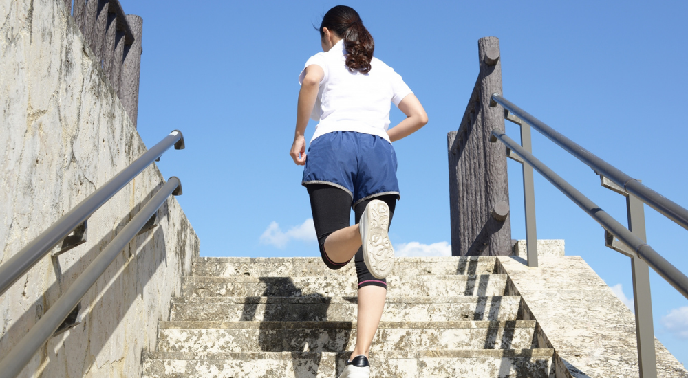 青空の下で女性が軽快に石段を駆け上がっている後ろ姿の画像で、【下半身・股関節(大臀筋・ハムストリングス周辺)】を示し、適切な身体の使い方や筋膜の滑走性向上により、足取りが軽くアクティブに運動を楽しめている様子。