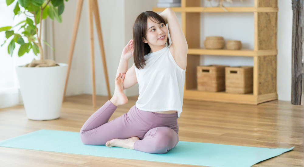 明るい部屋でヨガマットに座り、脚を曲げて腕を後頭部に回して体側や前ももを伸ばすポーズをとる女性の画像で、【股関節・体幹(腸腰筋・大腿四頭筋・広背筋周辺)】を示し、ヨガやストレッチによって縮こまった筋膜が心地よく引き伸ばされ、全身の柔軟性と血流が改善している様子。