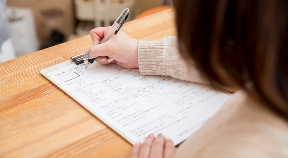 女性が、受付で、ご自身の【全身】の不調や悩みを【問診票】に記入している状況です。早く辛さを解消したいという真剣な思いと、これから体が良くなることへの期待を持って、ご自身の状態と向き合っている様子です。
