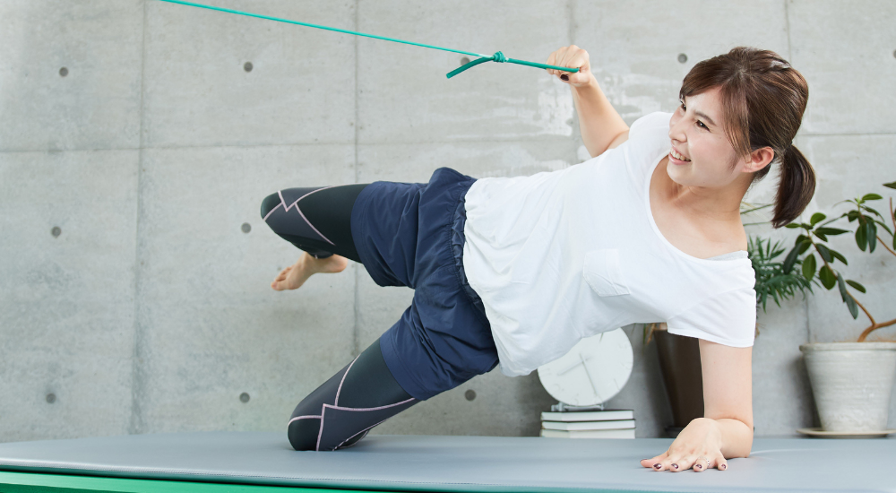 若い女性が、横向きで姿勢を保持しながらチューブを引き、【肩甲骨】を鍛えつつ、【体幹】や【中殿筋】などを鍛えるトレーニングを行っている状況です。キツい姿勢の中でも笑顔を見せ、前向きに楽しくご自身の体づくりに取り組んでいるアクティブな場面です。